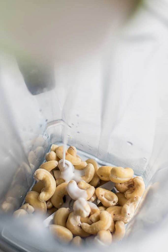 Cashews in a blender with coconut milk being drizzled in.