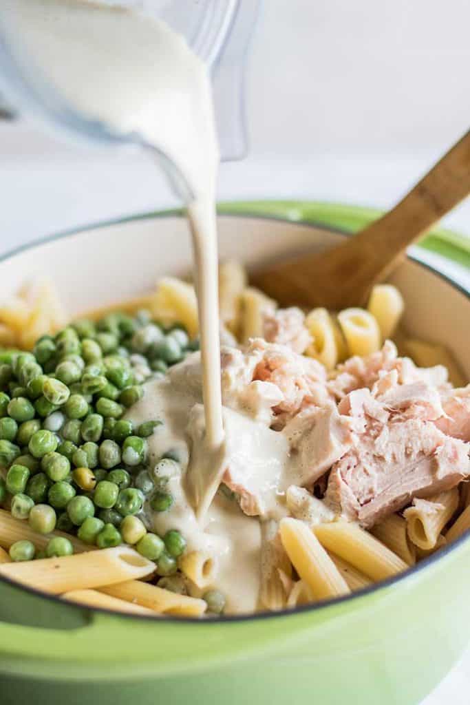 A pot of pasta with peas and tuna on top, with a dairy free cream sauce being poured in.