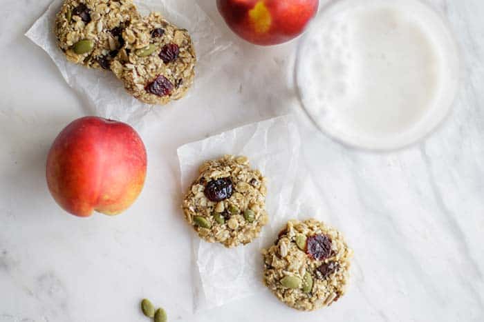 Several banana breakfast cookies on a table with glasses of milk and some fruit.