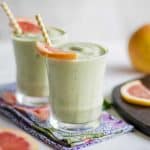 Two glasses filled with a green grapefruit smoothie, garnished with slices of grapefruit.