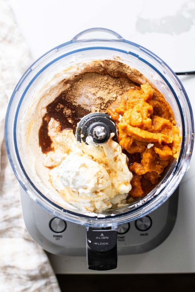 A food processor bowl shown with pumpkin, Greek yogurt, spices, and a sweetener added into the softened cream cheese.