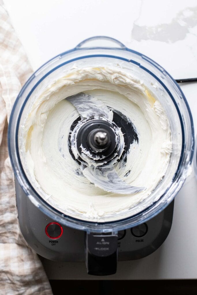Cream cheese in a food processor bowl shown being softened.