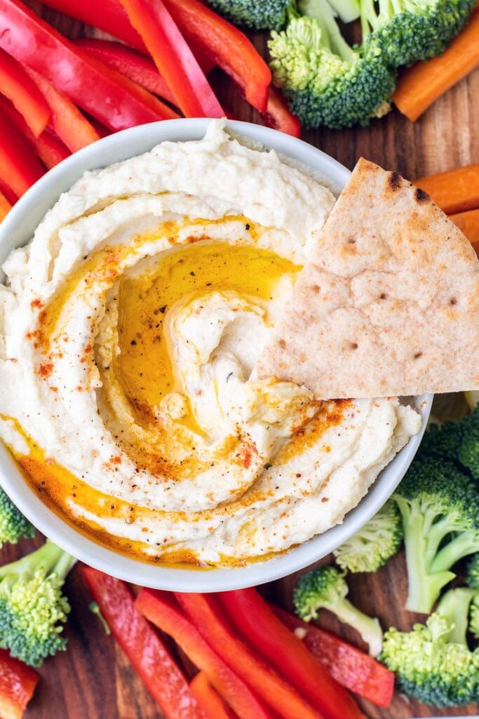 A bowl of hummus with colorful veggies and a pita wedge dipping some out.