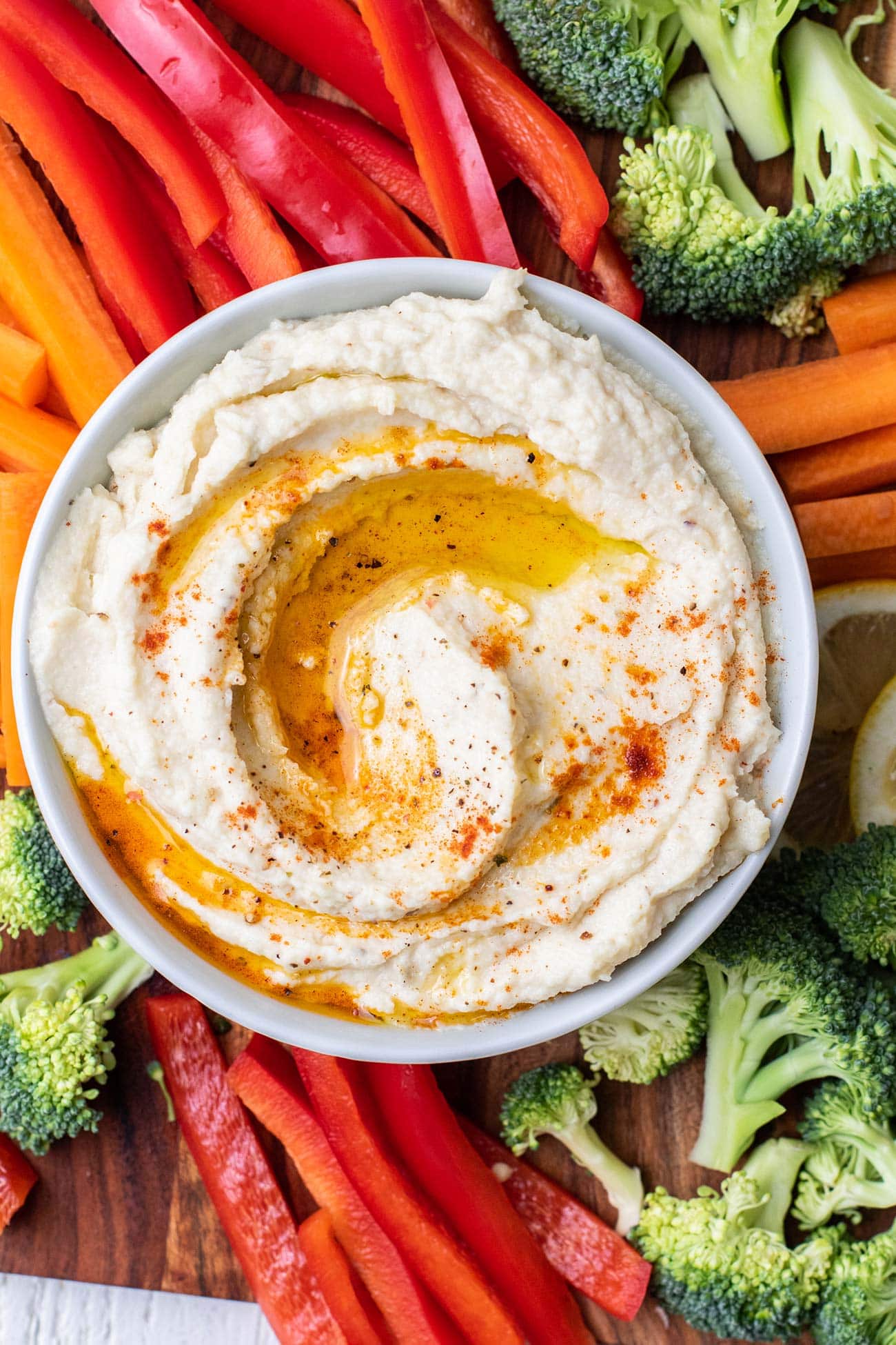 A bowl of cauliflower hummus shown surrounded by colorful veggies.
