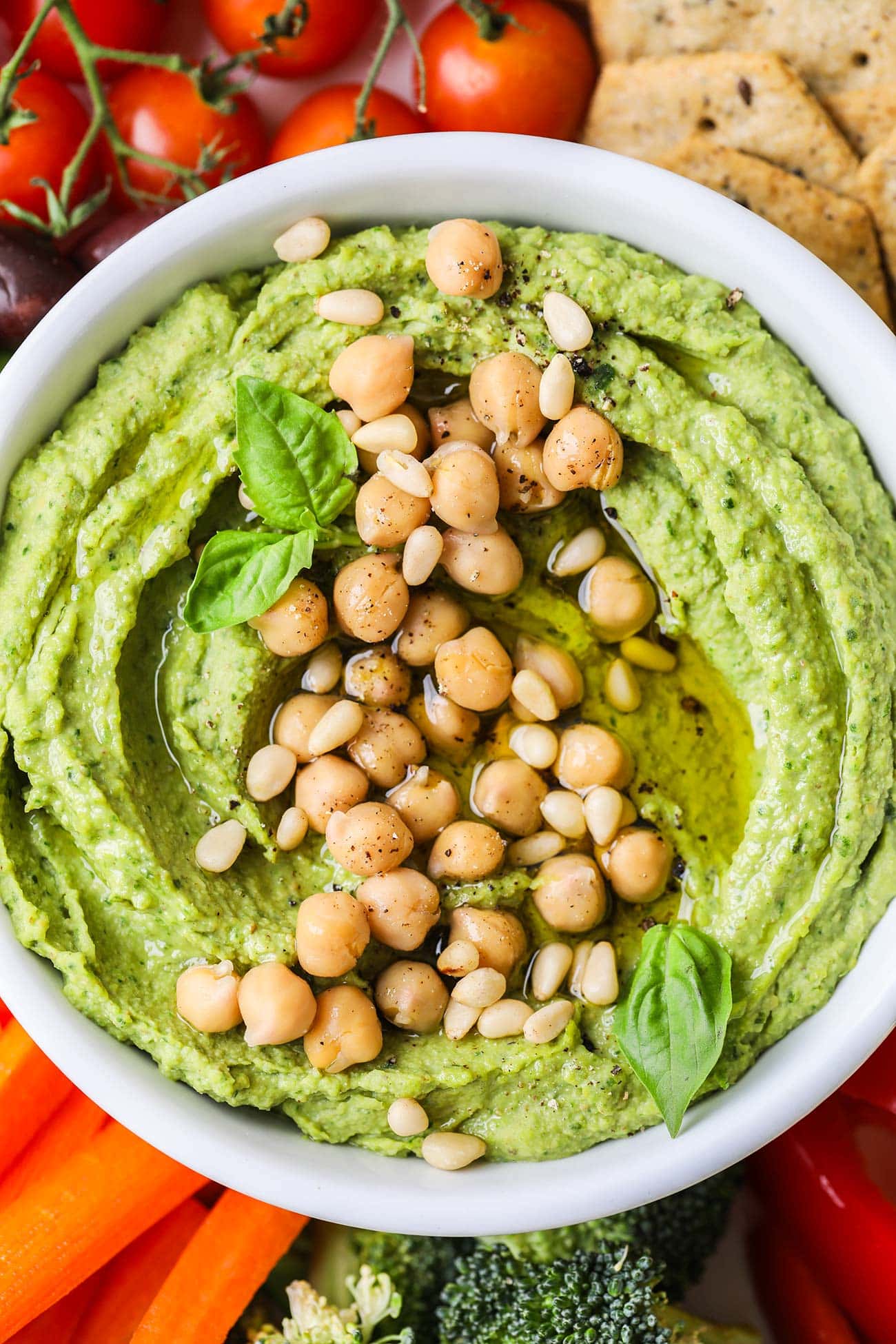 A vibrant green pesto hummus in a bowl surrounded by colorful veggies.