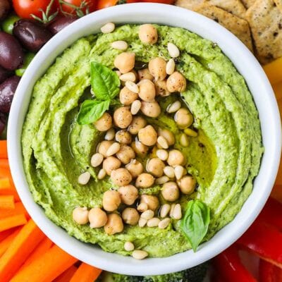 A bowl of bright green pesto hummus on a platter with colorful vegetables.