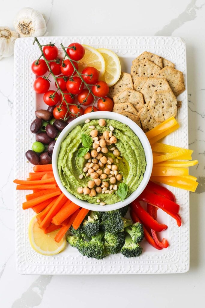 A large platter with colorful cut veggies and crackers, with a bowl of bright green pesto hummus in the center.