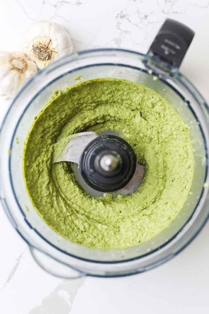 A food processor bowl with a vibrant green pesto hummus.