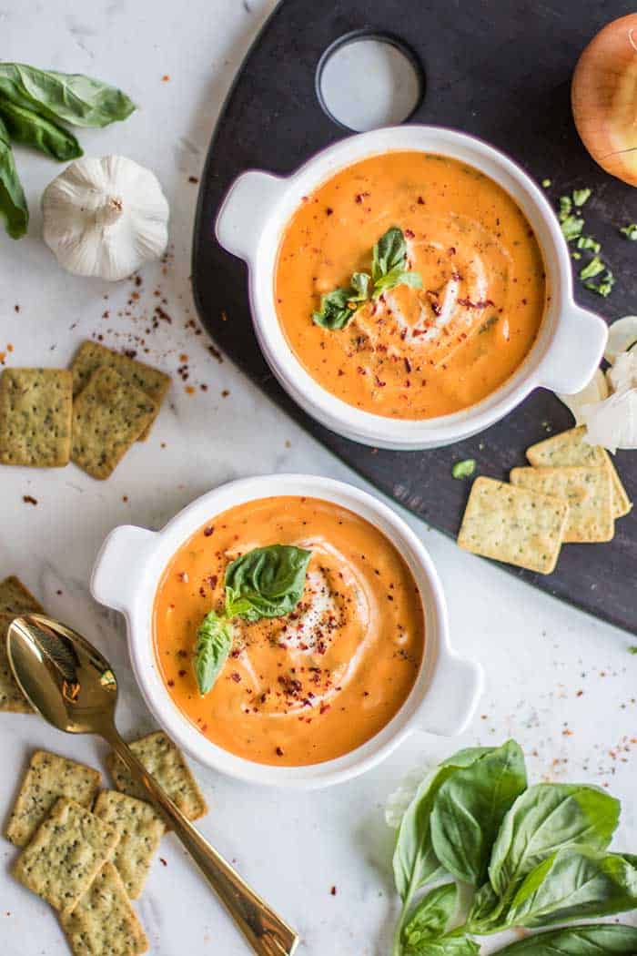 Two bowls of creamy tomato basil soup surrounded by whole grain crackers.