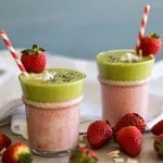 A layered Strawberry Colada green smoothie garnished with a strawberry and coconut and a red paper straw.