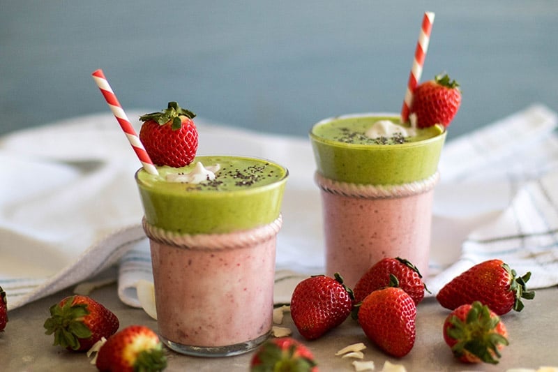 A layered Strawberry Colada green smoothie garnished with a strawberry and coconut and a red paper straw.