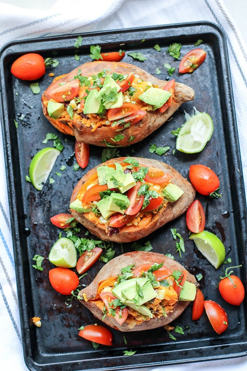 Baked sweet potatoes on a black baking tray topped with avocado, tomatoes, and cilantro.