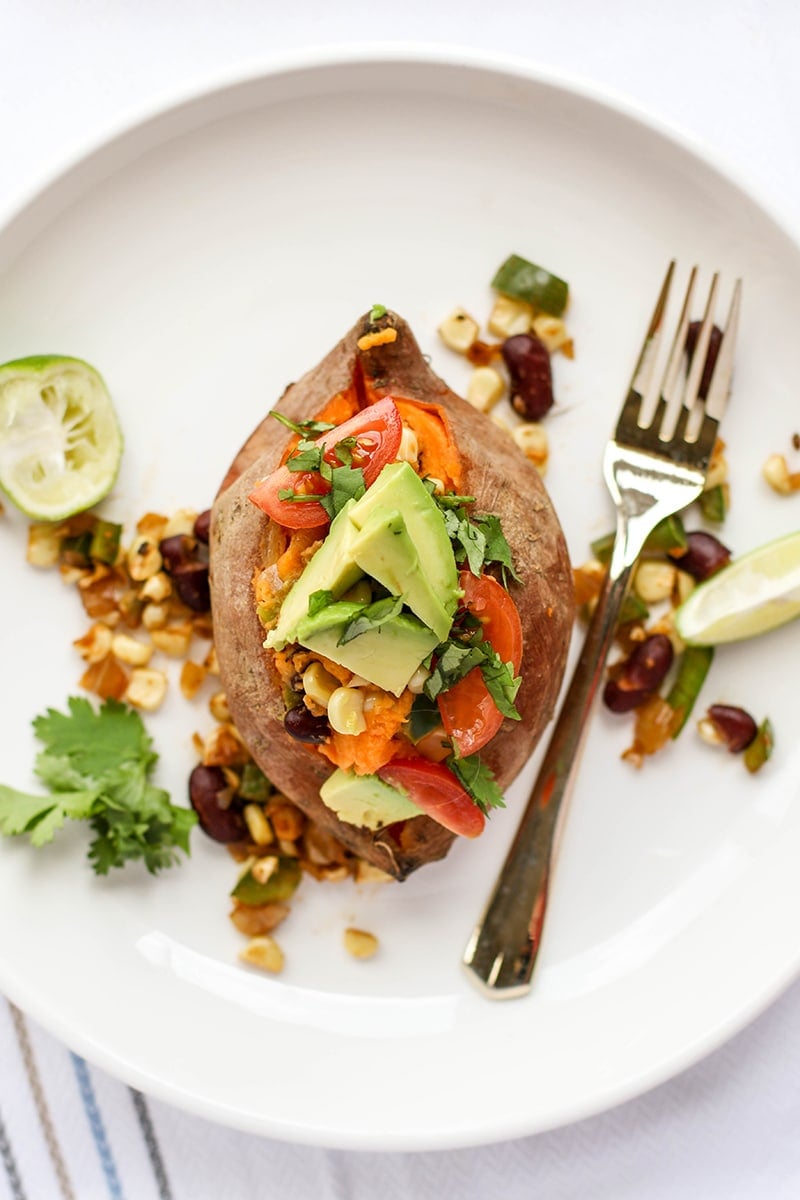 A stuffed sweet potato topped with avocado, cilantro, tomatoes and lime wedges.