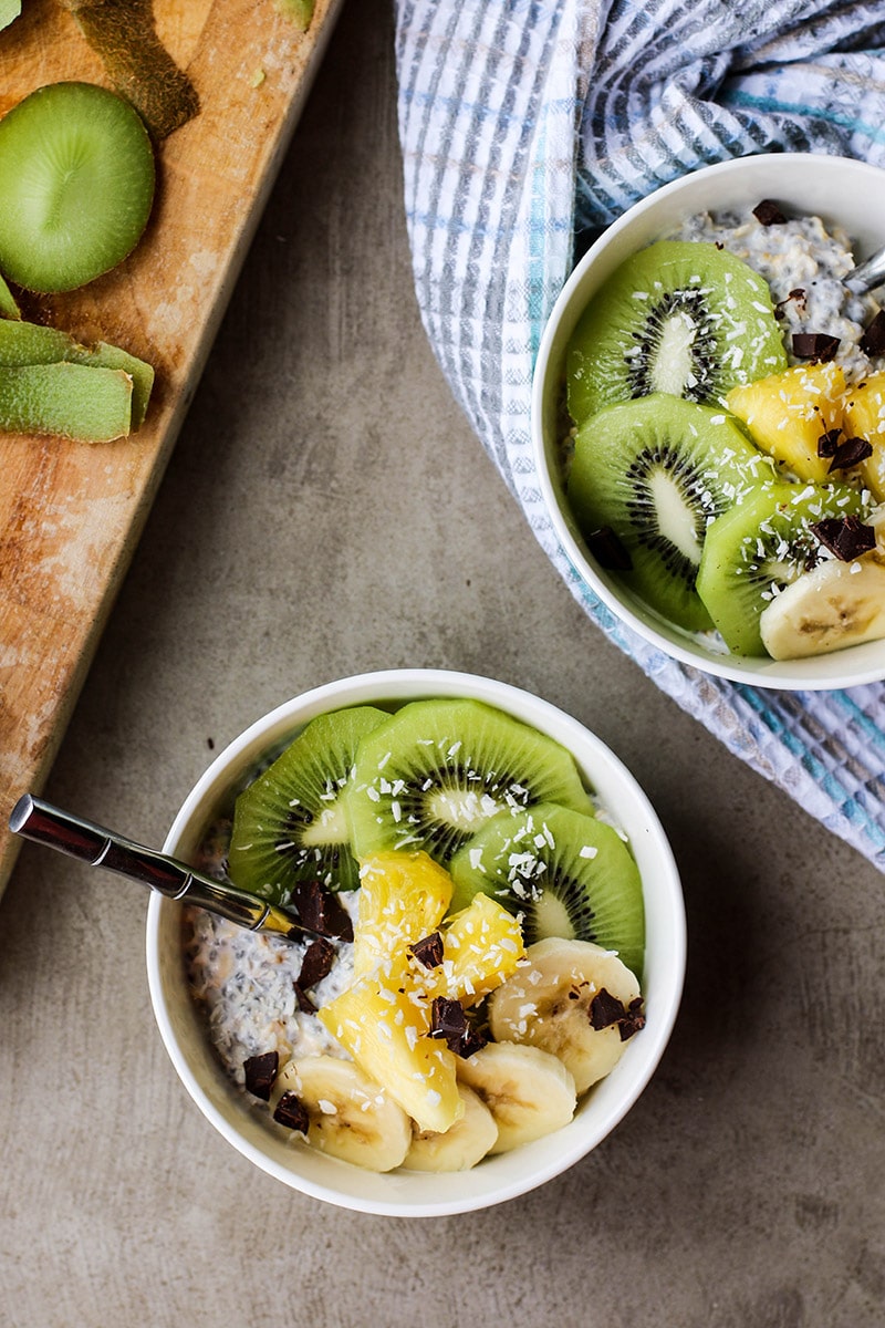 Two bowls of overnight oats with greek yogurt topped with kiwi, pineapple, bananas, and coconut.