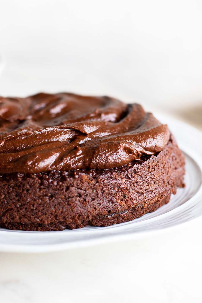 A chocolate quinoa cake on a white plate with a thick layer of fudgy frosting.