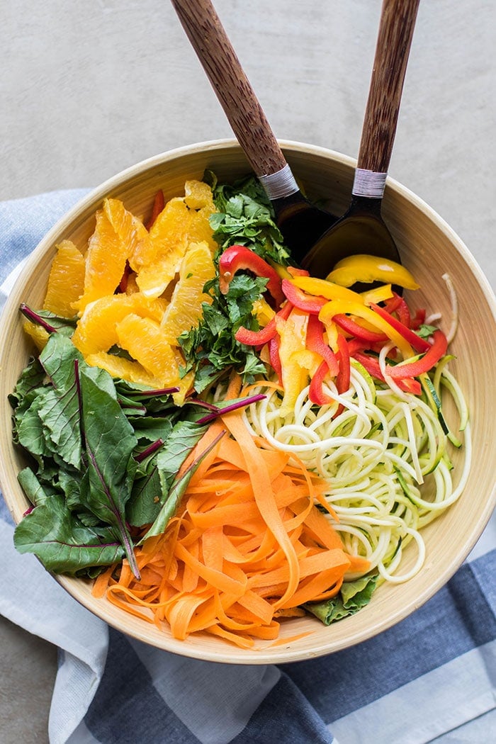 A bowl showing a colorful blend of zucchini noodles, carrot ribbons, thinly cut peppers, baby chard, and oranges.