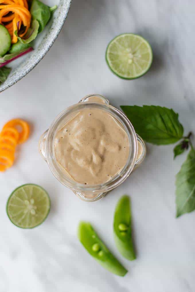 A spicy thai peanut sauce with almond butter shown in a jar with limes and cilantro.