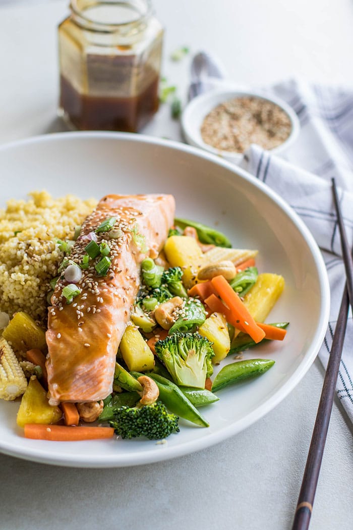 A plate with a piece of teriyaki salmon on top of a bed of millet and sauteed veggies.