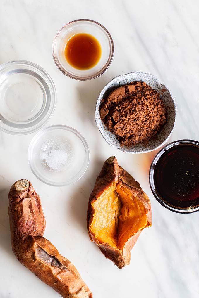 The ingredients for healthy chocolate frosting prepared and ready, including baked sweet potatoes, cocout oil, cocoa powder, date syrup, and vanilla.