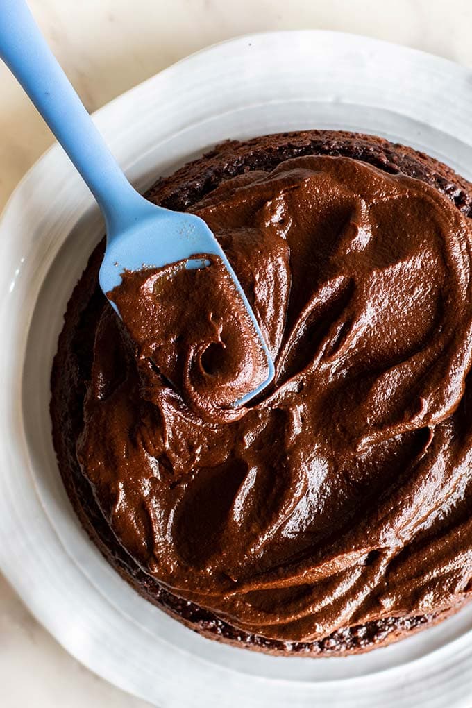 A blue spatula spreading a thick chocolate icing on a cake.