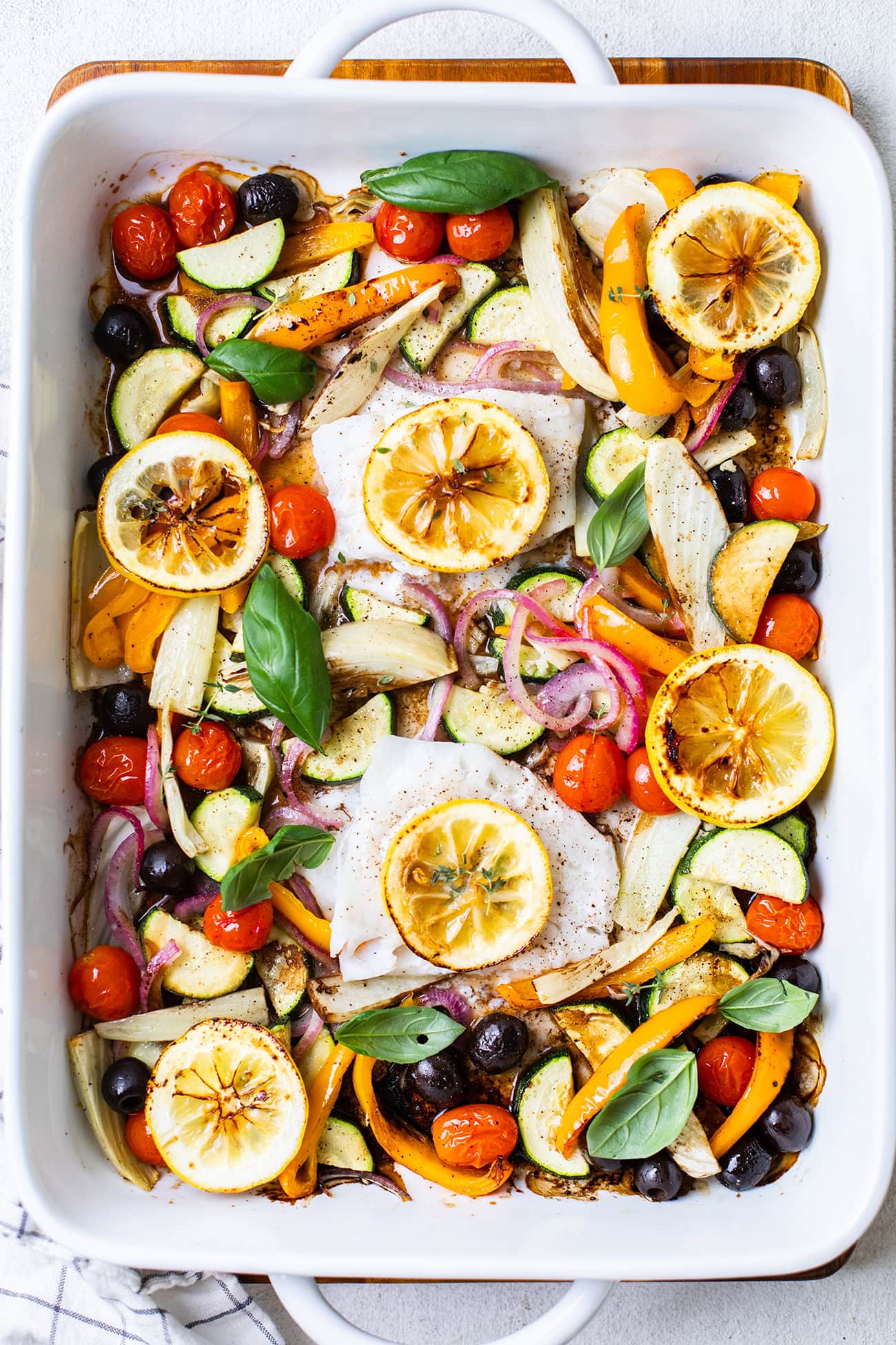 A large baking dish shown with 2 pieces of cod topped with lemon slices surrounded by colorful roasted vegetables.