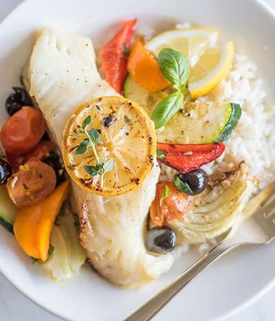 A piece of baked cod topped with a lemon slice, on a plate with rice and mediterranean vegetables.