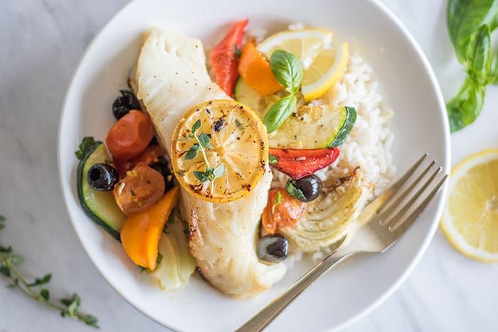 A piece of baked cod topped with a lemon slice, on a plate with rice and mediterranean vegetables.