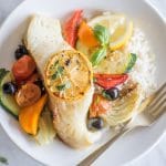 A piece of baked cod topped with a lemon slice, on a plate with rice and mediterranean vegetables.