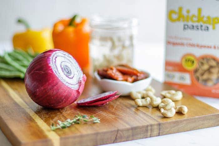 A cutting board with a box of gluten free pasta, and all the veggies being prepared for the dish.