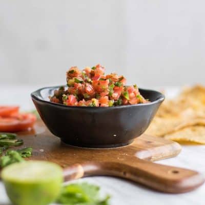 A cutting board showing a bowl of homemade salsa surrounded by fresh chopped ingredients.
