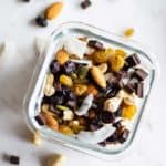 A close up of a container packed with trail mix, including nuts, coconut flakes, dark chocolate, and tart cherries.