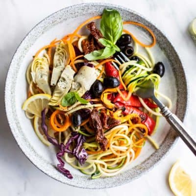 A plate with a large Italian Pesto Zoodle Salad, with some zucchini noodles twisted around a fork.