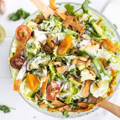 A clear bowl filled with a creamy Mexican caesar salad, with limes and cilantro around the bowl.