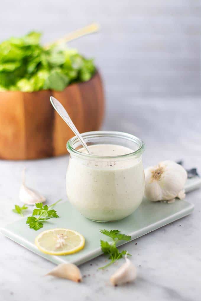 A jar of caesar dressing in front of a bowl of greens.