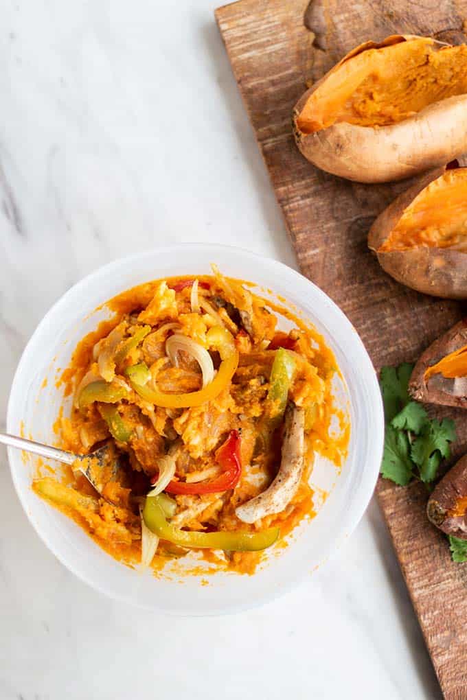 A bowl of mashed sweet potato with roasted chicken, onions and peppers mixed in, with hollowed out sweet potatoes.