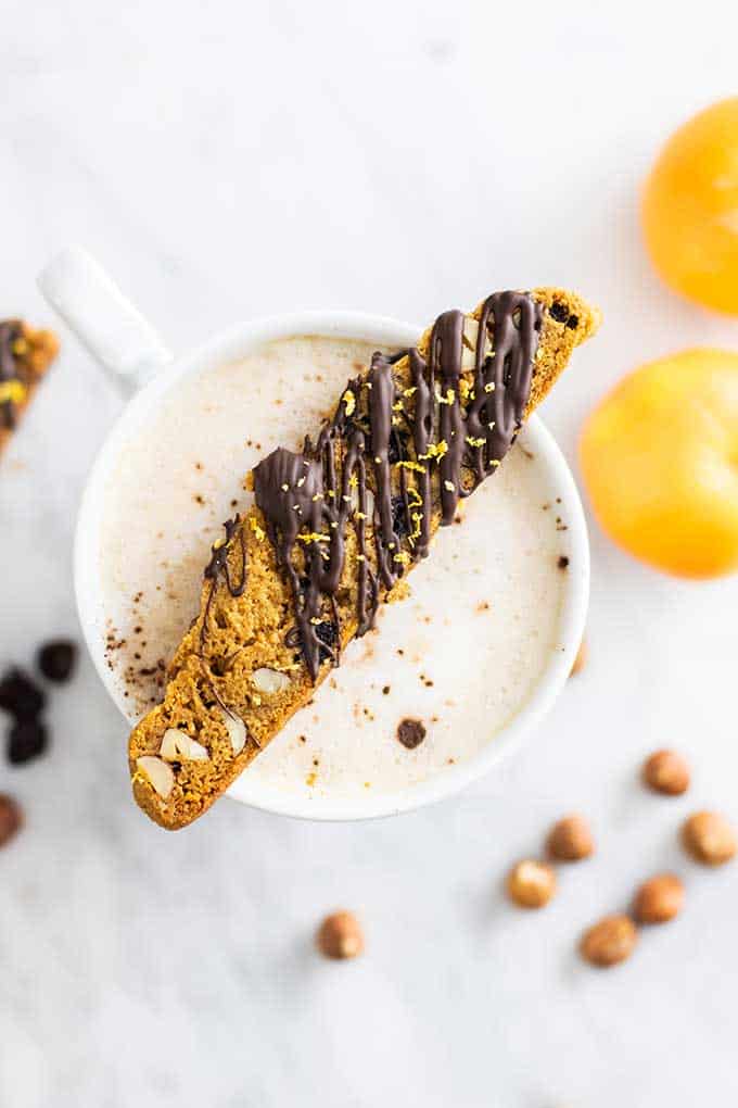 A slice of gluten free biscotti with a chocolate drizzle resting on top of a cappuccino.