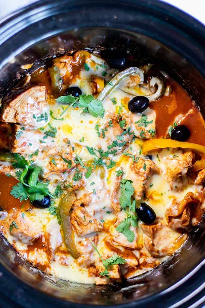 A crockpot filled with chicken, enchilada sauce, peppers, olives, and cilantro.