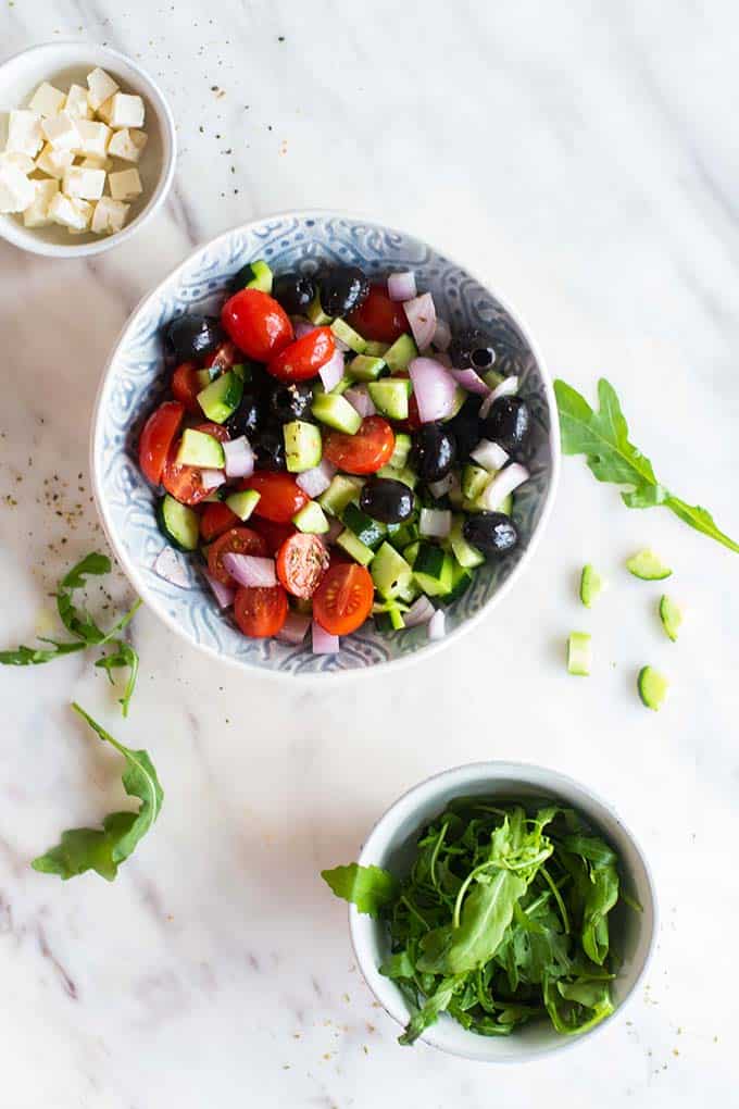 A bowl of greek salad, with arugula and feta cheese on the side.