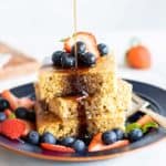 A stack of pancake squares topped with berries, being drizzled with maple syrup.