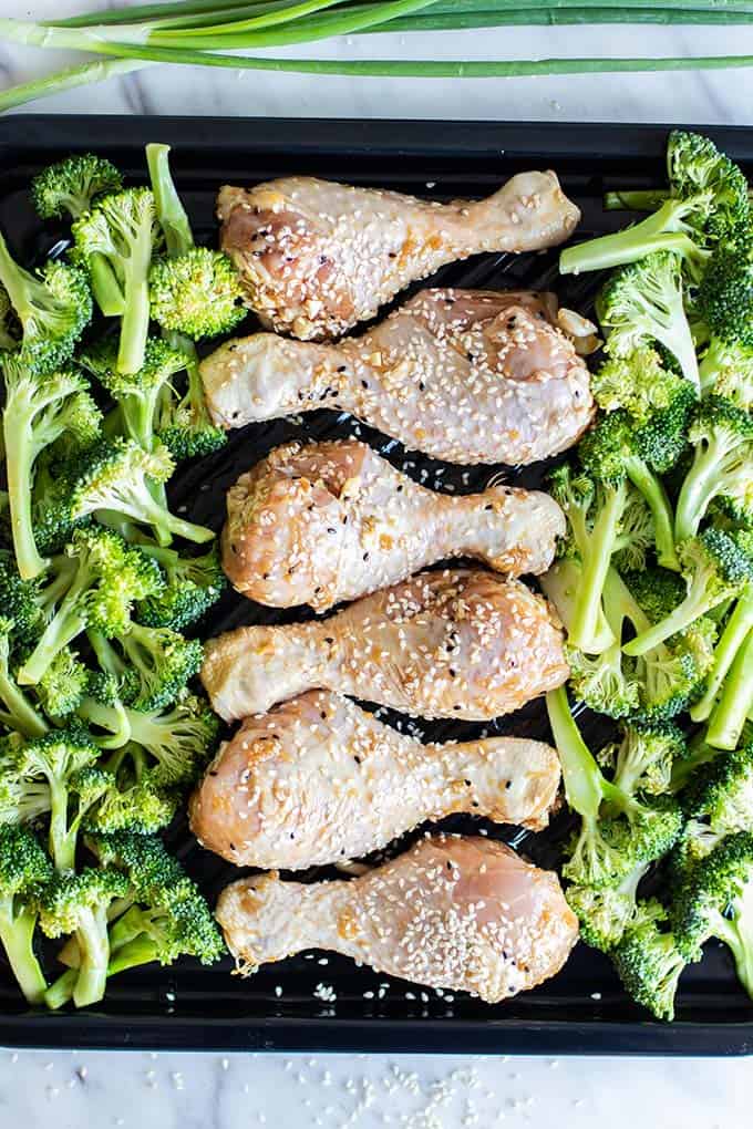 Raw marinated chicken legs on a baking pan with broccoli.