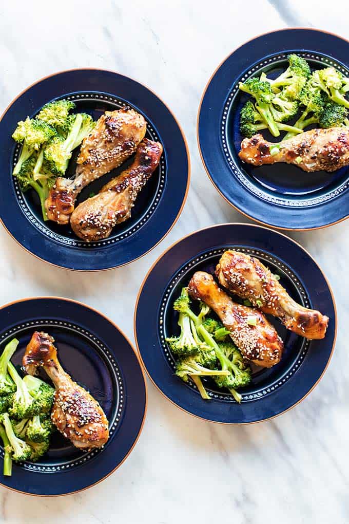 Four plates loaded up with roasted broccoli and baked sesame chicken legs.