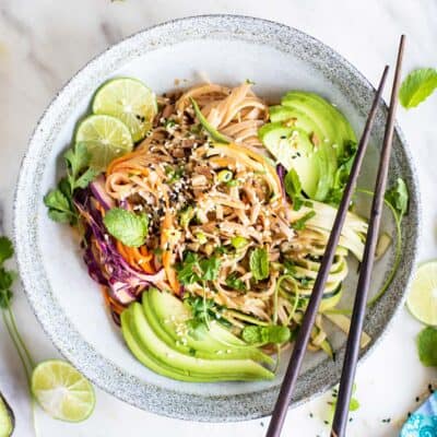 A spicy thai noodles dish garnished with avocado, sesame seeds and limes.
