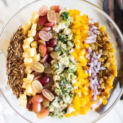 A large glass bowl filled with a broccoli cauliflower salad, with rows of toppings on top.