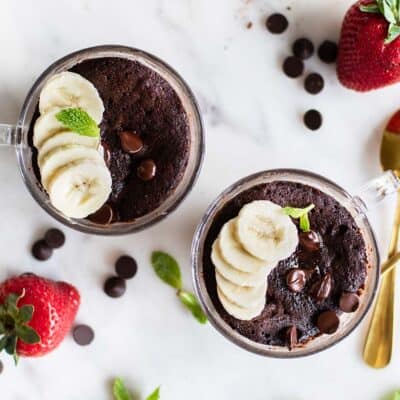 Two chocolate mug cakes in clear glass mugs, with chocolate chips, strawberries, and bananas.