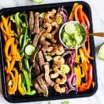A top view of a sheet pan with colorful peppers, onions, steak and shrimp, tossed in a fajita seasoning.