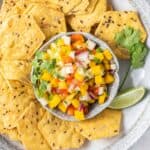 A bowl of multi grain chips with a dish or mango salsa garnished with limes and cilantro.