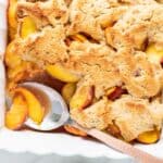 A baking dish filled with a golden brown peach cobbler with one scoop taken out.