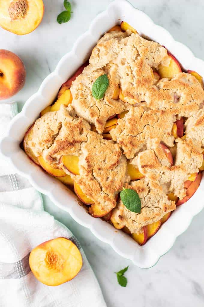A baking dish of peach cobbler with peaches and a white towel next to it.