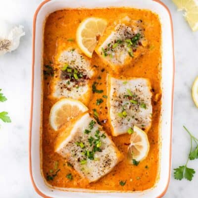 A baking dish with baked cod sitting in a creamy red pepper sauce.