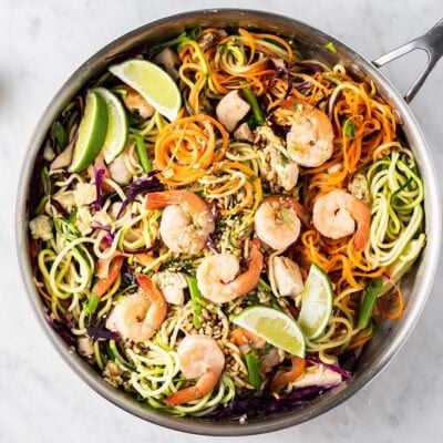 A large skillet filled with veggie noodles, chicken and shrimp, garnished with sunflower seeds and limes.